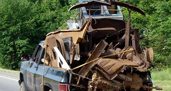 Tow truck transporting a heavily damaged scrap car on a forested roadside, highlighting trusted junk car buying in Rockwall, TX.